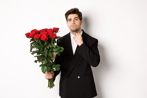 Immagine di un uomo elegante e sfacciato in abito nero, che sembra sicuro di sé e tiene in mano un mazzo di rose rosse, che va ad un appuntamento romantico, in piedi su sfondo bianco.