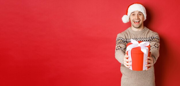 Immagine di un uomo felice che dice buon Natale e ti dà un regalo in una scatola sorridente allegro che celebra le vacanze invernali con cappello da Babbo Natale e maglione di Natale in piedi su sfondo rosso