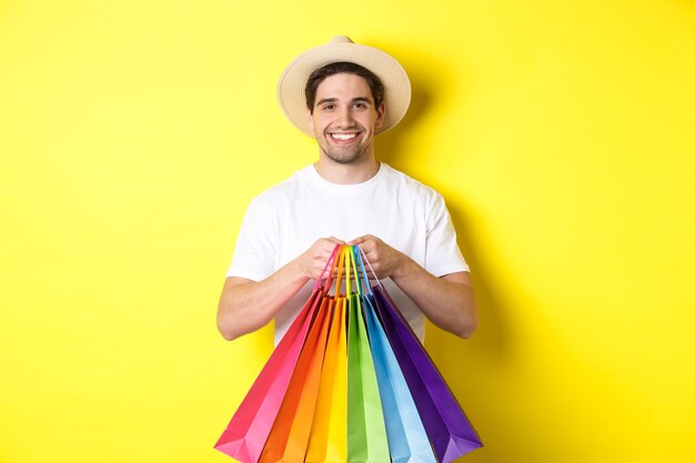 Immagine di un uomo felice che fa shopping in vacanza, tenendo in mano i sacchetti di carta e sorridente, in piedi su sfondo giallo.