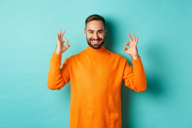 Immagine di un uomo sorridente fiducioso che mostra il segno giusto, approva e accetta, garantisce la qualità, in piedi sul muro turchese chiaro.