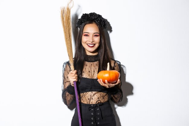 Immagine di una bella donna asiatica vestita come una strega per la festa di Halloween, tenendo scopa e zucca, in piedi su sfondo bianco.