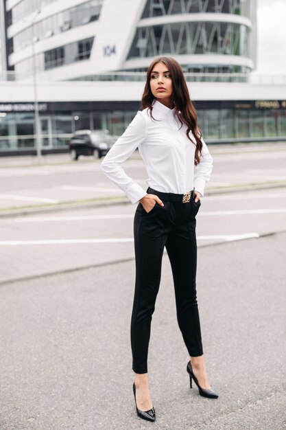 Immagine di una bella donna caucasica con lunghi capelli mossi scuri in una camicia bianca, pantaloni neri e tacchi in posa per la fotocamera