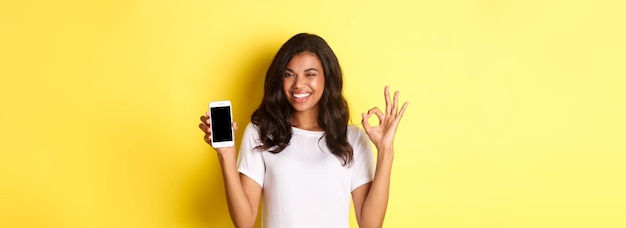 Immagine di una bella ragazza afroamericana che strizza l'occhio e sorride mostrando lo schermo dello smartphone e il segno ok