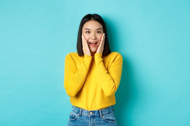 Immagine di una donna asiatica allegra e sorpresa che controlla la promozione, ansimando stupita, in piedi su sfondo blu
