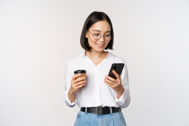 Immagine di una donna asiatica moderna che guarda il telefono cellulare che beve caffè da asporto con gli occhiali utilizzando l'app per smartphone in piedi su sfondo bianco