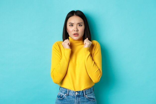 Immagine di una donna asiatica preoccupata con i capelli corti e scuri, stringe le mani e fissa la telecamera interessata, in piedi su sfondo blu