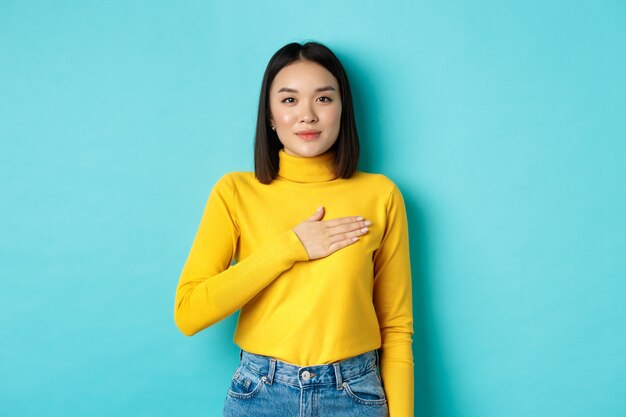 Immagine di una donna asiatica sorridente orgogliosa che tiene la mano sul cuore, mostrando rispetto per l'inno nazionale, in piedi su sfondo blu