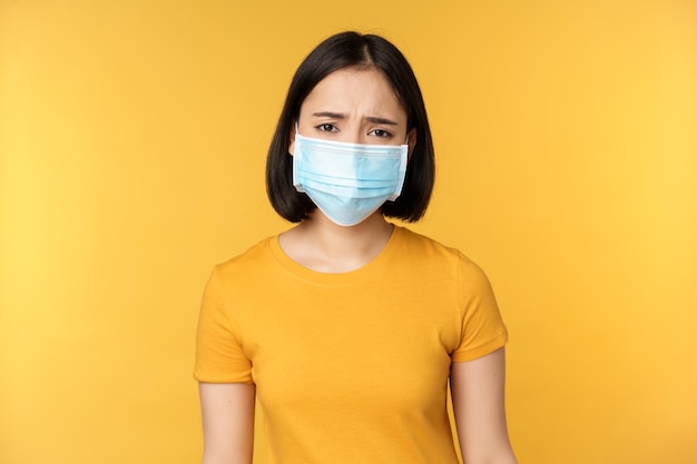 Immagine di una donna asiatica triste e delusa in maschera medica accigliata e che sembra scontenta in piedi su sfondo giallo
