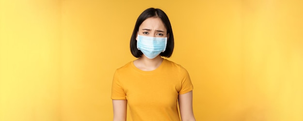Immagine di una donna asiatica triste e delusa in maschera medica accigliata e che sembra scontenta in piedi su sfondo giallo