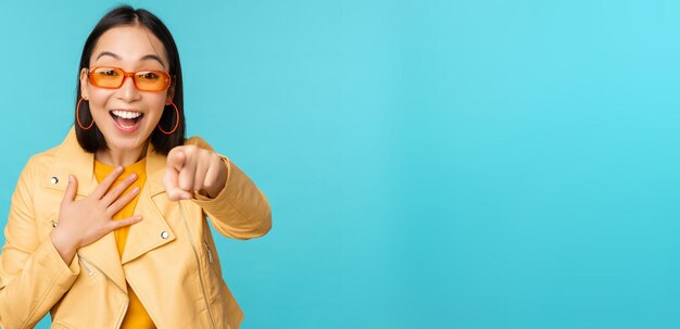 Immagine di una donna coreana felice in occhiali da sole che punta il dito verso la fotocamera con un'espressione del viso sorpresa e gioiosa stupita in piedi su sfondo blu Spazio di copia