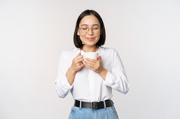 Immagine di una donna coreana sorridente che annusa l'odore del caffè nella tazza godendo di una bevanda deliziosa in piedi su sfondo bianco