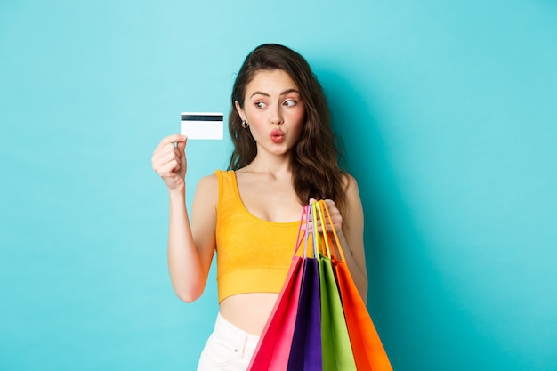 Immagine di una donna felice dello shopping che mostra la sua carta di credito in plastica, tiene in mano le borse della spesa, indossa abiti estivi, in piedi su sfondo blu.