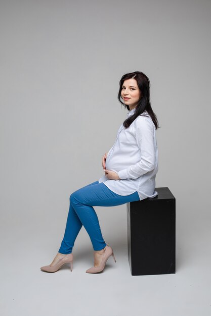 Immagine di una donna incinta allegra con capelli neri e bel sorriso in camicia bianca e jeans blu seduto su una sedia e in posa per la fotocamera