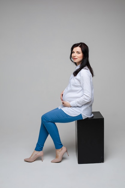 Immagine di una donna incinta allegra con capelli neri e bel sorriso in camicia bianca e jeans blu seduto su una sedia e in posa per la fotocamera