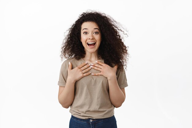 Immagine di una donna sorpresa ed eccitata che vince, riceve notizie fantastiche e gioisce, guarda in soggezione con un sorriso bianco felice, tenendo le mani sul petto stupita dall'annuncio, sfondo bianco