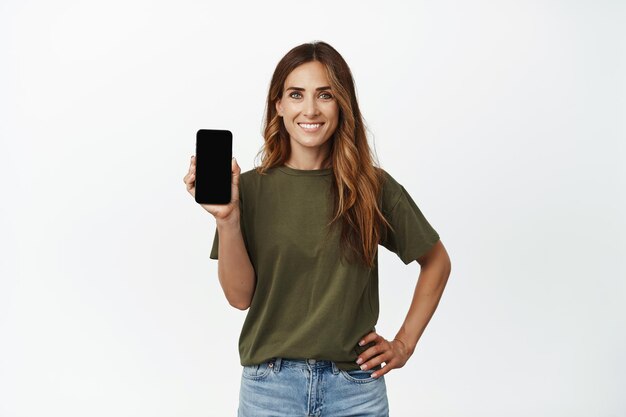 Immagine di una donna sorridente ed elegante che mostra lo schermo, il display mobile vuoto, l'interfaccia del telefono, l'app consigliata, la pubblicità cellulare 4g, in piedi su sfondo bianco.