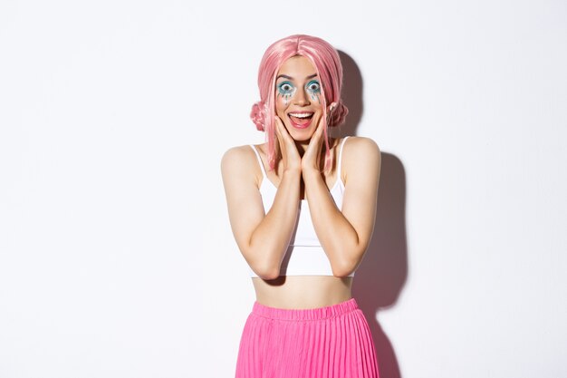 Immagine di una festaiola eccitata con parrucca rosa e trucco luminoso che sembra impressionata, sorridente e che guarda sorpresa, in piedi.