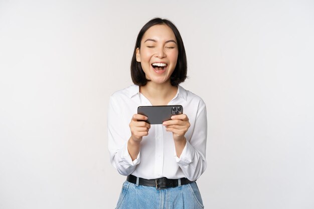 Immagine di una giovane donna asiatica che guarda sull'app per smartphone con in mano il telefono cellulare e guarda lo schermo ridendo e sorridendo in piedi su sfondo bianco