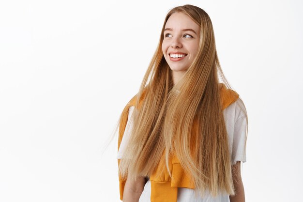 Immagine di una giovane donna attraente con lunghi capelli lisci naturali e sani che volano in aria, gira la testa e guarda a sinistra il testo promozionale, sorride felice, in piedi su sfondo bianco