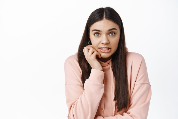 Immagine di una giovane donna bruna imbarazzante che si china e sembra colpevole, commettendo un piccolo errore sciocco, in piedi su un muro bianco