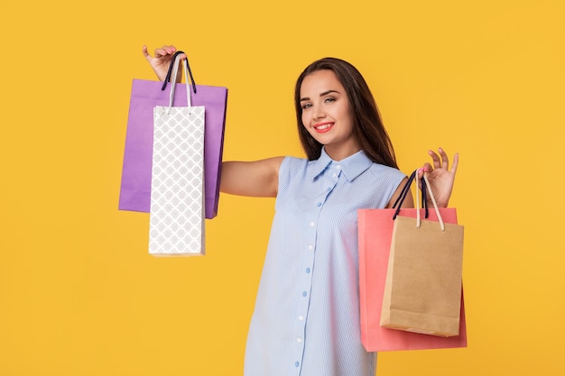 Immagine di una giovane donna bruna in abito a righe bianco e blu estivo in posa con borse della spesa e guardando la fotocamera su sfondo giallo. Shopping. Vendita