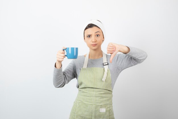 Immagine di una giovane donna carina in grembiule con una tazza blu che mostra un pollice verso il basso