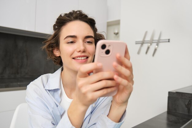 Immagine di una giovane donna che guarda il suo smartphone leggendo un messaggio seduta a casa e facendo acquisti online pagando le utilità su un'app mobile