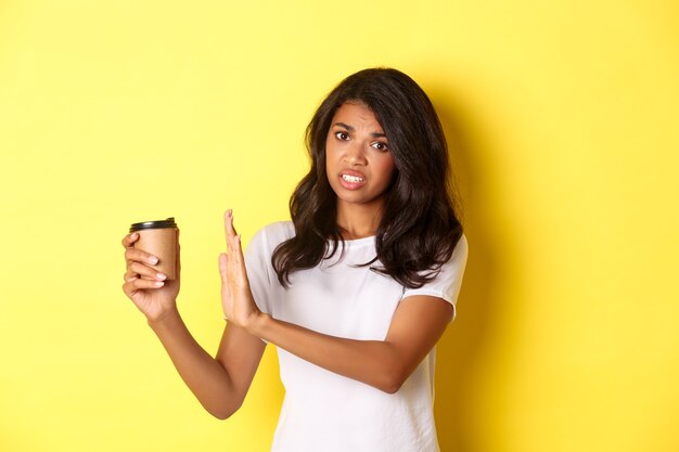 Immagine di una ragazza afro-americana che si lamenta del cattivo gusto del caffè, mostrando il segno di rifiuto e tirando via la tazza, in piedi su sfondo giallo.