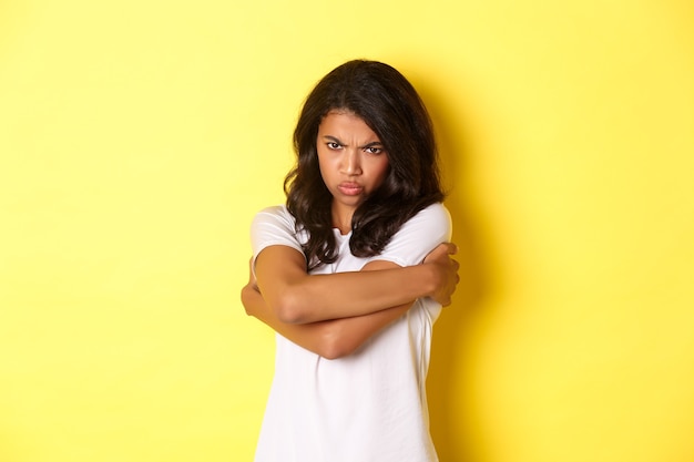 Immagine di una ragazza afroamericana carina e lunatica, che si abbraccia e tiene il broncio con un'espressione offesa, in piedi su sfondo giallo.