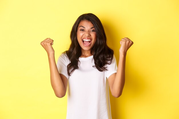 Immagine di una ragazza afroamericana di successo, che si sente fortunata, fa il segno della pompa a pugno e dice di sì, trionfa e grida di gioia, in piedi su sfondo giallo.