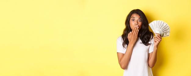 Immagine di una ragazza afroamericana sorpresa che vince soldi ansimando stupita in piedi su sfondo giallo