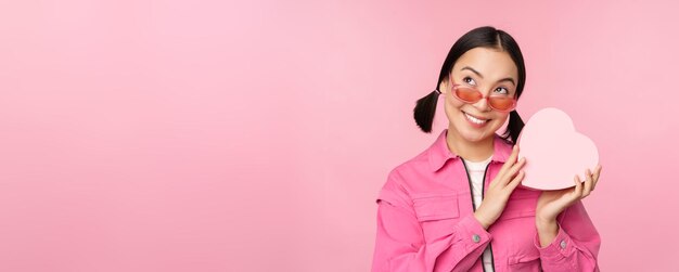 Immagine di una ragazza asiatica alla moda in occhiali da sole che indovina cosa c'è all'interno della confezione regalo presente a forma di cuore s