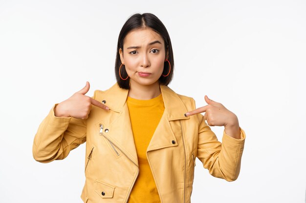 Immagine di una ragazza asiatica non divertita che punta a se stessa guardando con incredulità scettica alla telecamera in posa su sfondo bianco