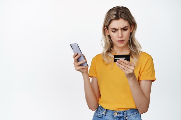 Immagine di una ragazza bionda seria che tiene il telefono cellulare guardando il numero di carta di credito con la faccia concentrata che cerca di pagare online in piedi su sfondo bianco