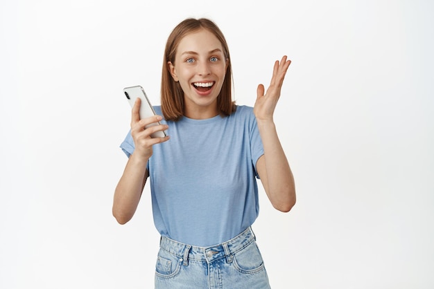 Immagine di una ragazza bionda sorpresa che salta dalla felicità grandi notizie sul telefono cellulare che tiene lo smartphone e si rallegra vincendo online in piedi su sfondo bianco