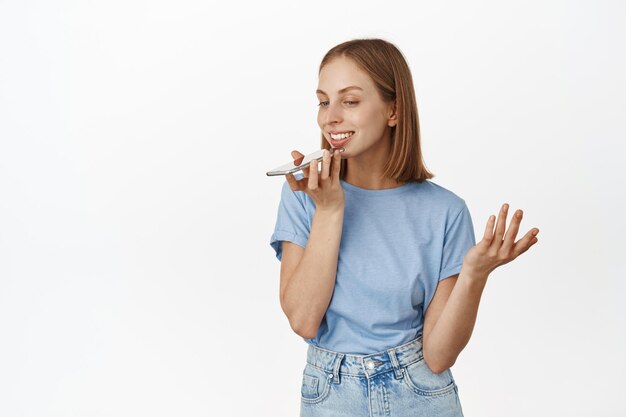 Immagine di una ragazza bionda sorridente che parla in vivavoce, telefono cellulare vicino alla bocca delle labbra, gesticola mentre parla, registra un messaggio vocale, traduce le sue parole nell'app mobile, sfondo bianco.