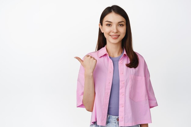 Immagine di una ragazza caucasica sorridente che punta a sinistra guardando la fotocamera che consiglia la vendita del negozio in piedi su sfondo bianco