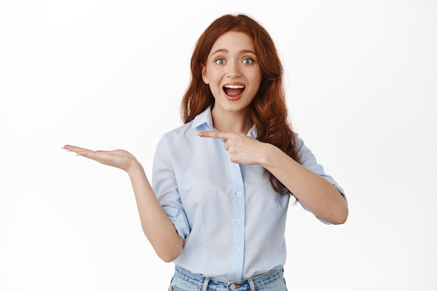 Immagine di una ragazza dai capelli rossi stupita che punta il dito verso la mano aperta, tenendo il tuo prodotto di marca e consigliandolo, mostrando l'oggetto in mano, in piedi felice su sfondo bianco