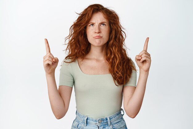 Immagine di una ragazza rossa imbronciata che punta le dita verso l'alto e sembra arrabbiata per qualcosa in piedi in maglietta su sfondo bianco