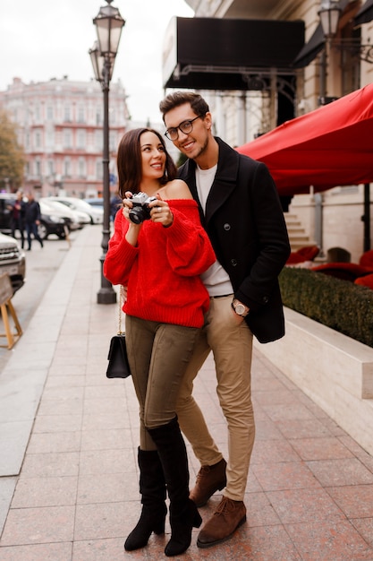 Immagine esterna a tutta altezza di coppia elegante alla moda innamorata che cammina per strada durante la data o le vacanze. Donna castana in maglione rosso che fa le foto dalla macchina fotografica.