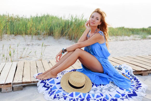 Immagine estiva all'aperto di donna romantica in cappello di paglia rilassante sulla spiaggia assolata in abito blu.