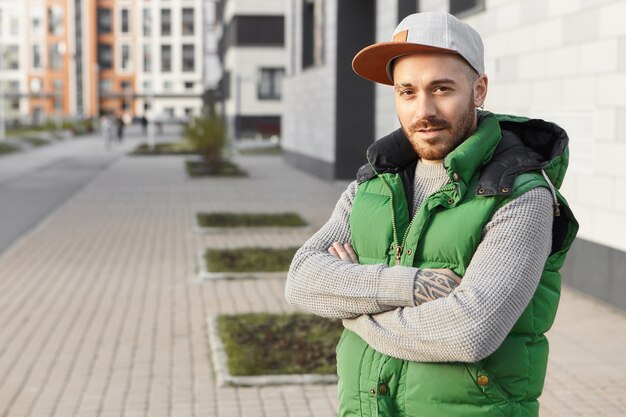 Immagine estiva orizzontale di muscoloso giovane maschio atletico vestito con abiti eleganti, incrociando le braccia sul petto, in posa all'aperto, andando a fare una corsa mattutina. Stile, fiducia e concetto di moda