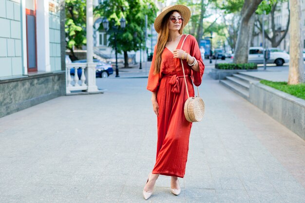 Immagine integrale della donna alla moda che trascorre le sue vacanze in città europea. Indossa un fantastico abito boho di corallo alla moda, tacchi, borsa di paglia.