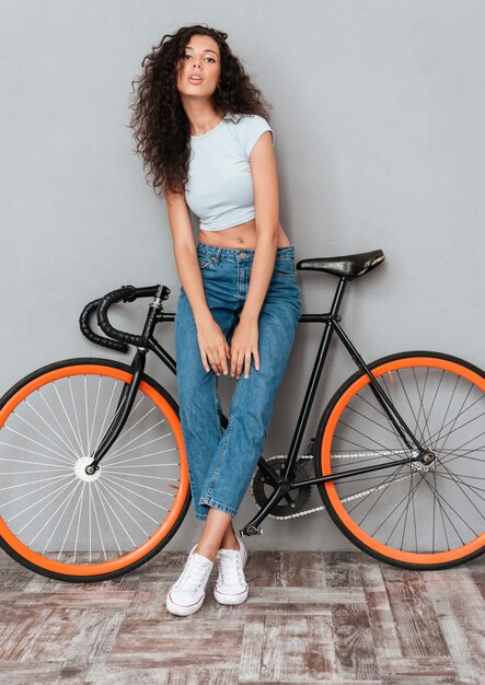 Immagine integrale della donna graziosa riccia che posa con la bicicletta e che esamina la macchina fotografica sopra fondo grigio