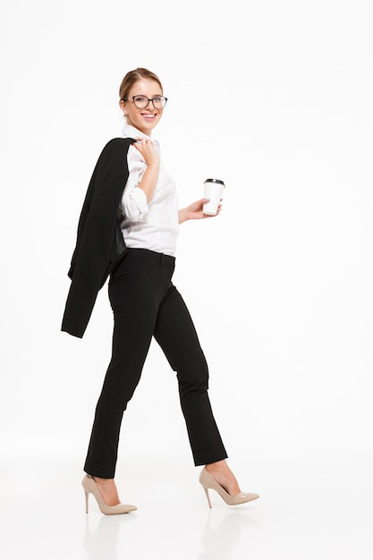 Immagine integrale di vista laterale della donna bionda sorridente di affari in occhiali che cammina nello studio con la tazza di caffè e sopra la parete bianca
