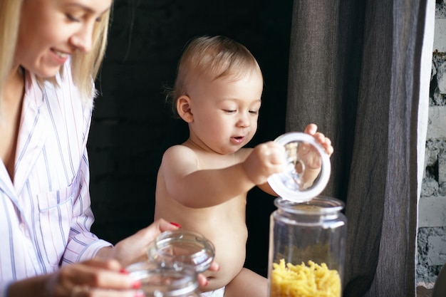 Immagine interna di adorabile bambino felice in pannolino che gioca con il coperchio della bottiglia di vetro in cucina, la sua bella giovane madre seduta accanto a lui, sorridendo ampiamente. Messa a fuoco selettiva sul viso del bambino