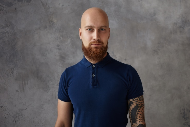 Foto gratuito immagine isolata del ragazzo calvo con folta barba rossa e tatuaggio mentre posa contro il muro bianco con copia spazio per il tuo testo, informazioni o contenuto promozionale