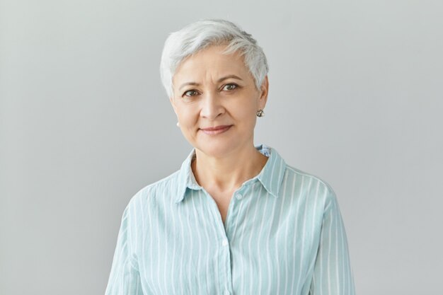 Immagine isolata di amichevole bella donna d'affari europea con i capelli grigi pixie sorridendo fiduciosamente, felice con i risultati del lavoro del suo team, vestito in camicia a righe formale