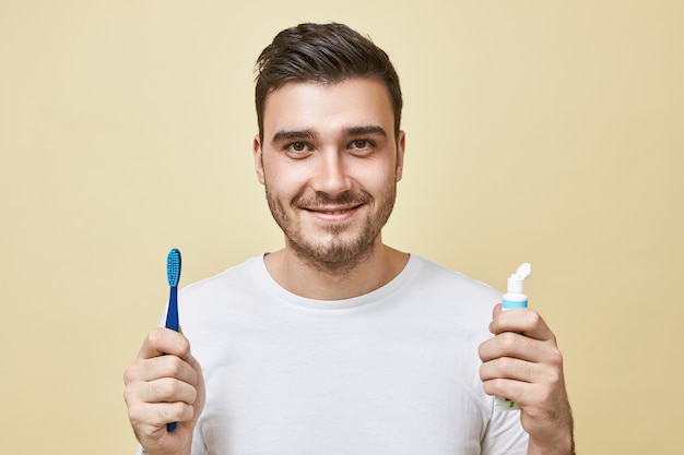 Immagine isolata di fiducioso allegro giovane brunetta ragazzo con setole che tiene pennello e tubetto di dentifricio, lavarsi i denti subito dopo il risveglio. Igiene, routine mattutina e concetto di sbiancamento dei denti