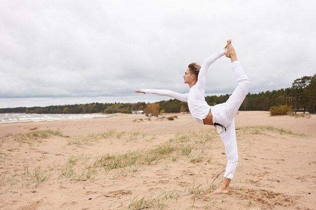 Immagine laterale di un ragazzo attraente in piedi a piedi nudi con un piede sulla sabbia, facendo Natarajasana o la posa di King Dancer sulla spiaggia deserta. Giovane maschio a praticare Hatha yoga all'aperto in riva al mare